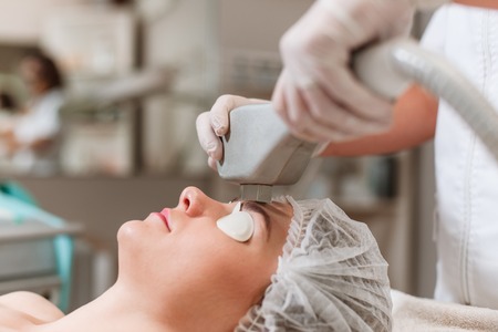 Portrait of young woman receiving laser treatment by a doctor-cosmetologist in special protective glasses. Removing pigmentation at cosmetological clinic. Anti-aging procedures and laser hair removal.の写真素材