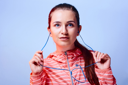 Young happy girl in bright jacket with hood and headphones listening to the music and dancing isolated on blue background.の写真素材