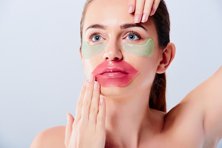 Portrait of beauty woman with natural make-up applying blue hyaluronic hydrogel patches and pink lips mask. Female face with mask under eyes. Skin care concept. Korean cosmetics. Isolated on greyの写真素材