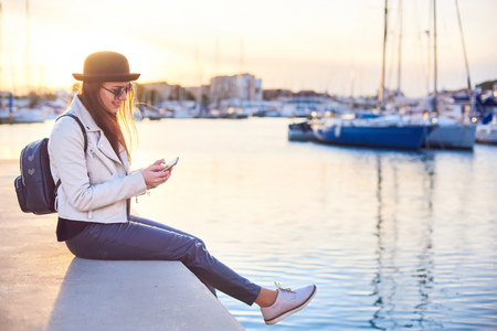 Pretty young long-haired woman blogger wearing black hat and round sunglasses is smiling while sitting on a pier and texting on a cell phone at the sunset in a port with sea and boats on background.の写真素材