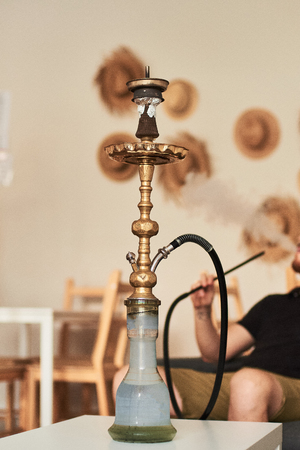 Close-up of hookah with red coconut coal and man smoking shisha at lounge bar on background.