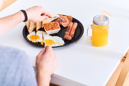 Man eating traditional full English breakfast with fried eggs, sausages, beans, bacon and toasts sitting at white table.の写真素材