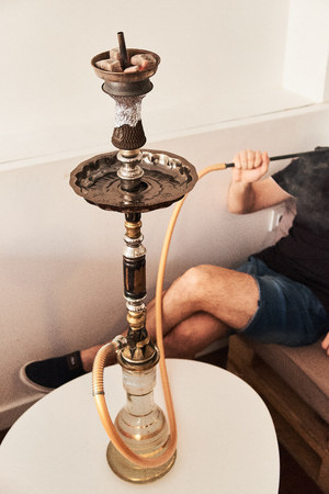 Close-up of hookah with red coconut coal and man smoking shisha at lounge bar on background.