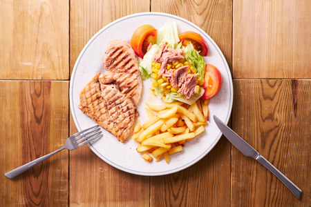 Overhead shot of French fries with grilled meat and tuna salad. Plate on wooden background.の写真素材