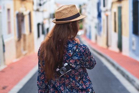 Back view of a young long-haired woman in hat walking down the street with small colorful buildings.の写真素材