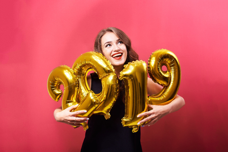 Young smiling woman with bright make-up celebrating New Year 2019 and holding golden balloons in hands on red background.の写真素材