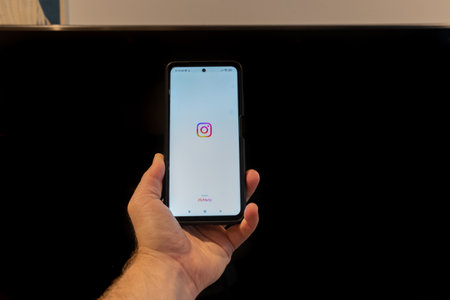 a hand hold up an illustration of the instagram logo displayed on a smartphone screen , in front of a tvのeditorial素材