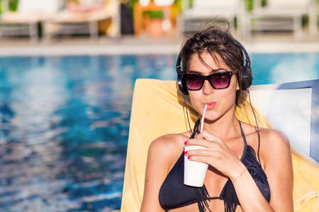 Portrait of beautiful young woman listening music and drinking juice on the poolの写真素材