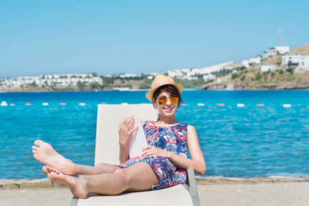 Portrait of beautiful young woman sitting on a sunbed with tablet in the hands on a sea background.Summer holidayの写真素材