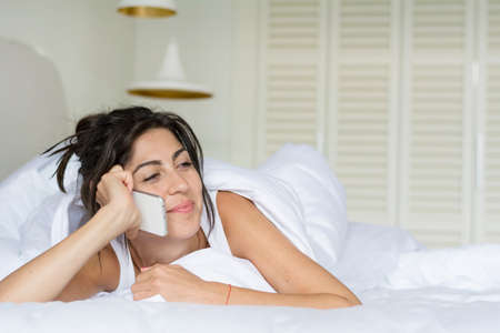 beautiful young woman on a white linen in bed at home,talking on the phoneの写真素材