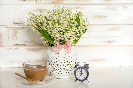 Bouquet of lily-of-the-valley flowers in vase, alarm clock and cup of coffee on white wooden backgroundの写真素材