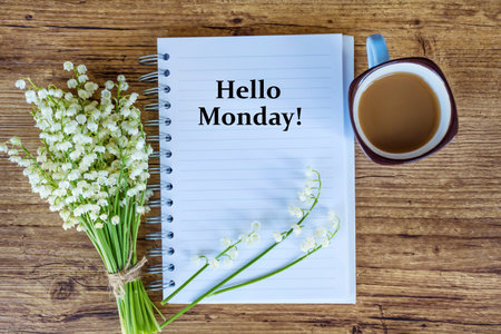 Lily of the valley, cup of coffee and notebook with text Hello Monday on wooden backgroundの写真素材
