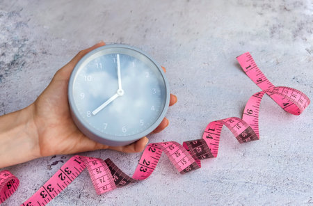 Time for diet and weight loss. Female hand holding clock and measuring tapele background.