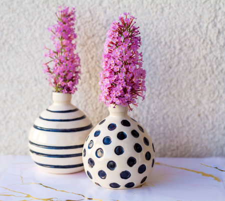 Two ceramic vases with purple flowers on a white table against a white wallの写真素材