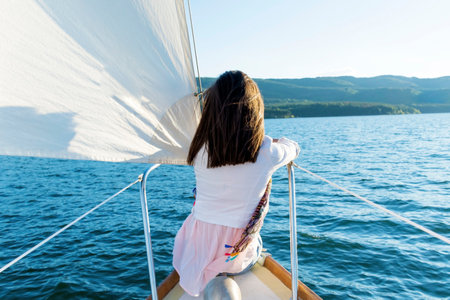 Rear view of a young girl standing on the deck of a sailboat and looking at the lakeの写真素材