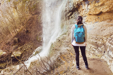 Young woman with backpack standing in front of waterfall and looking at itの写真素材