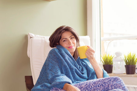 Portrait of a young woman wrapped in a blanket drinking coffee at home.の写真素材