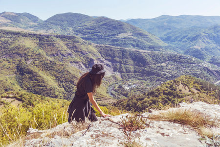 Young woman in black dress and hat standing on top of a mountain and looking at the valleyの写真素材