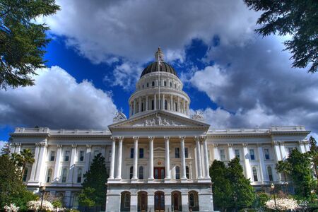 an HDR image of the California State Capital Building from a distance bordered by trees and a blue sky with grey and white cloudsの写真素材
