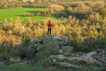 a man in a red jacket and with a backpack stands on top of a stone block and looks at the sunlit wooded valleyの写真素材