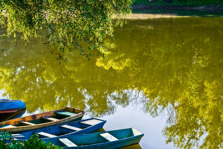 Quiet sunny morning on the bank of a small river. A group of boats are moored to the shore under the green willow branches. Light steam can be seen above the mirror-like waterの写真素材