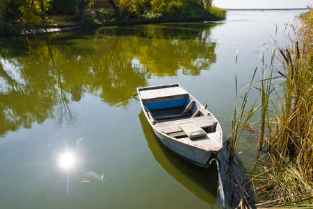 The fishing boat is moored to the shore of the river bay next to the reed mace thickets. Trees and sun are reflected in the water. Farm Obukhovka, Rostov region, Russiaの写真素材