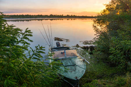 Summer dawn on the Don River near the village of Bagaevskaya. An old metal boat with a motor is moored to the shore overgrown with bushes. The first rays of the sun break through the foliage of the treesの写真素材