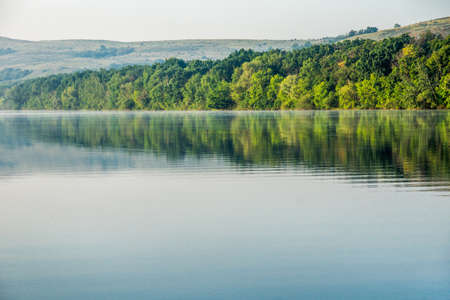 Quiet summer morning on the Seversky Donets River near the town of Belaya Kalitva, Russia. A light haze spreads over the mirror-like water of the river. In the water reflections of the coastal forestの写真素材