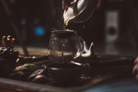 pouring tea from ceramics teapot into glass tea bowl on bamboo tea tray, warm dark atmosphere during tea ceremonyの写真素材