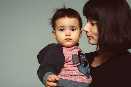 Profile of young mother with dark short hair holding her son toddler, serious sad child with chubby cheeks, looking at camera, isolated in studio, space for textの写真素材