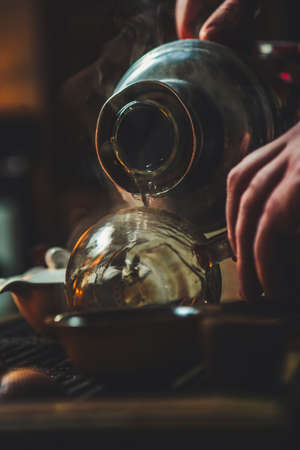 pouring tea from jug into glass jar on bamboo tea tray, warm dark atmosphere during tea ceremony