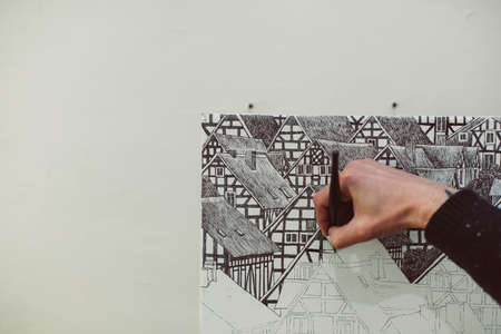 Male hand of artist is drawing with ink pen, monochrome picture of houses, white background, lots of...