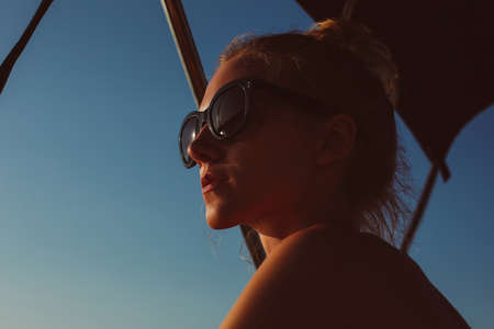 Profile portrait of young woman looking into the distance, wearing green sunglasses, relaxed face, fair hair in bun, blue skyの写真素材