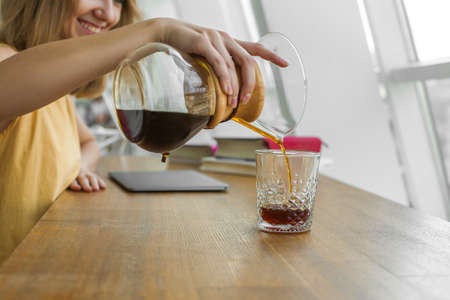 happy woman with graceful hand is pouring coffee from glass jar into glass, near window, wooden...