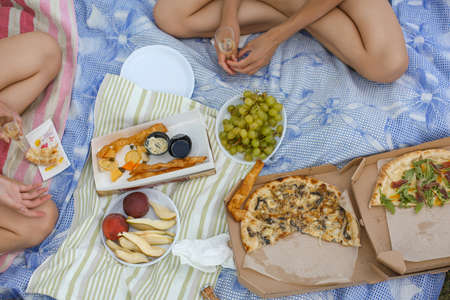 top view of two girls sitting cross-legged on the blue blanket, picnic, pizzas, fruits, glass with wineの写真素材