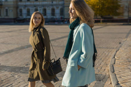 Two young women are speaking, walking in old city centre, wearing elegant coats, warm sunny dayの写真素材