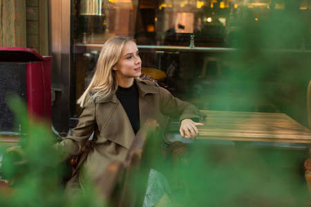 Stylish young woman with blond hair, wearing khaki coat, sitting at wooden table near window in antique cafeの写真素材