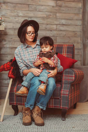 Portrait of young mother with small son sitting in checked armchair wearing checked shirts, hat, in decorated Scandinavian interiorの写真素材
