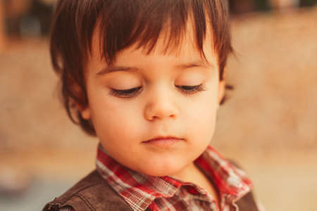 Close up warm portrait of the face of small boy with dark messy hair, long eyelashes, sweet beautiful face, almost closed eyes, looking down, checked collar shirtの写真素材