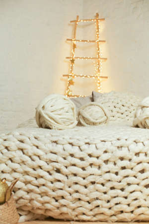 knitted white blanket on bed with decorative clew in light Scandinavian interior, wooden ladder with fairy lightsの写真素材