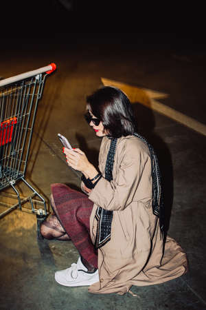 stylish fashionable young woman, using her phone, wearing beige trench coat, white sneakers, short black glossy hair, on hunkers, dark streetの写真素材