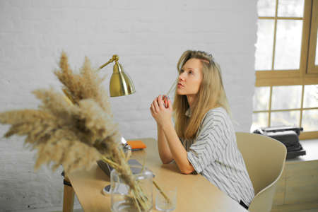 Beautiful concentrated young woman with blond hair, holding pencil, is thinking, wearing white striped shirt, sitting at wooden table, with lamp, decoration, white wall background, windowの写真素材