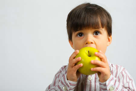 Small caucasian boy with dark hair, big dark eyes, wearing striped shirt is eating, biting big green apple, isolated on white backgroundの写真素材