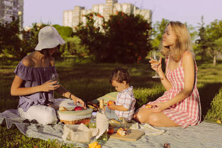 Two female friends and small child are sitting on the blanket on green grass, in park, drinking wine, having a picnicの写真素材
