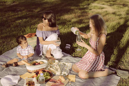 Two female friends and small child are sitting on the blanket on green grass, in park, drinking wine, having a fun. laughing, having picnicの写真素材