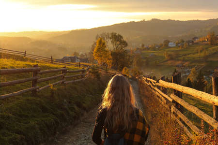 A girl in a plaid jacket walks along a country road in the mountains at sunset.の写真素材