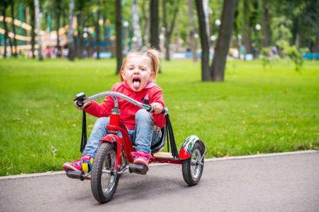Little girl in amusement park riding on tricycle on the road and showing tongue