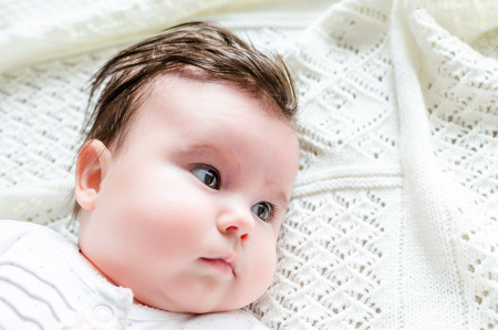 Portrait of cute sweet little newborn baby girl with black brunette hair in nice white spotted romper suit looking with big beautiful hazel brown eyes lying on white woolen blanket on a bed at homeの写真素材