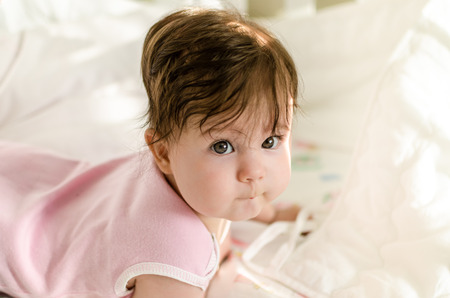 Cute sweet little baby girl with black brunette hair in nice pink romper suit lying on her bed at...