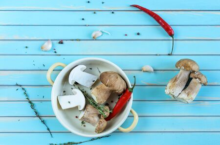 Wild fresh porcini mushrooms in metal frying pan served with fresh herbs, red hot chili peppers, garlic and thyme over stylized old aged wooden turquoise table background, top view. Rustic background with copy spaceの写真素材